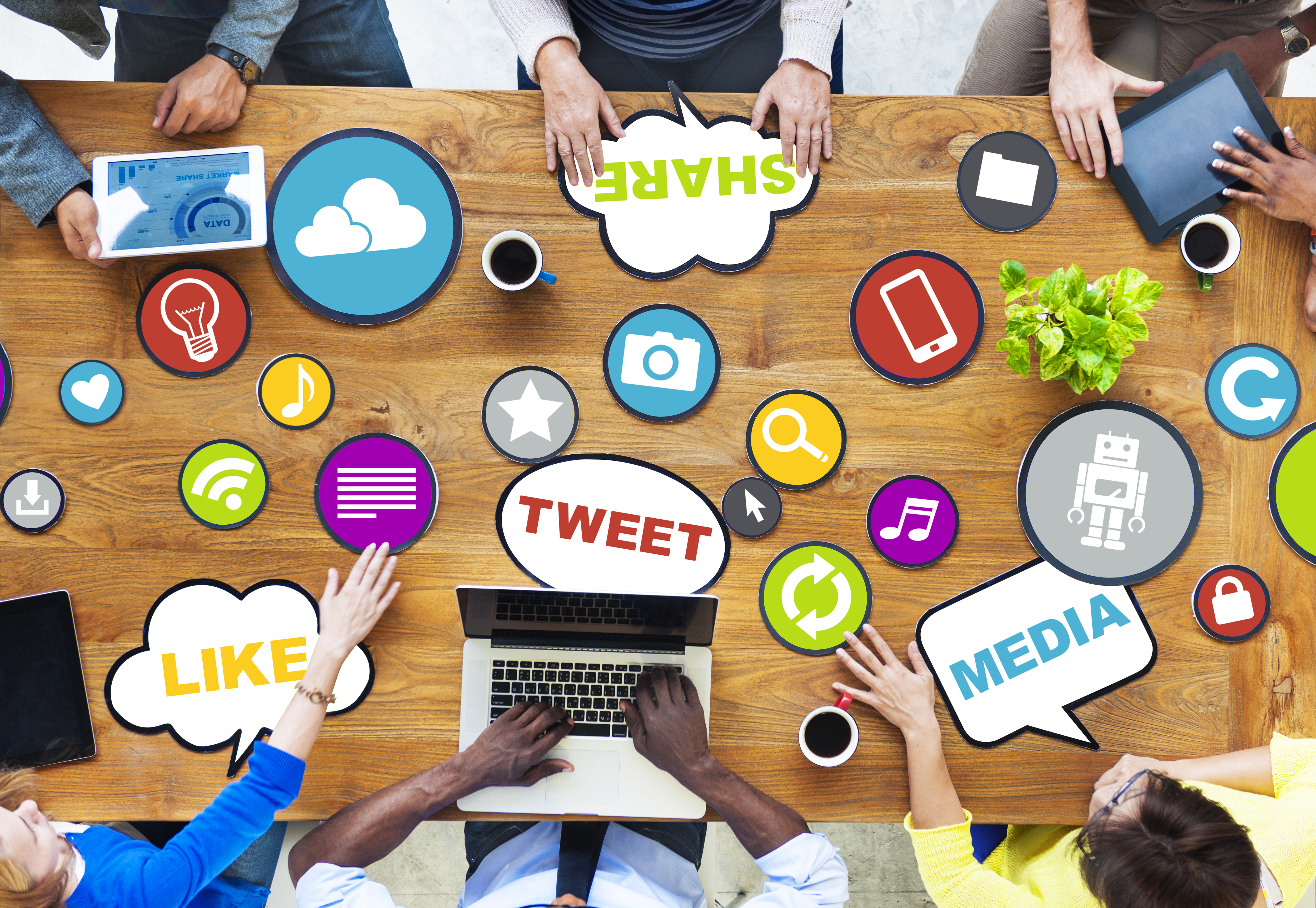 A meeting desk pictured from above. Several people are working on electronic devices and social media icons are scattered in front of them.